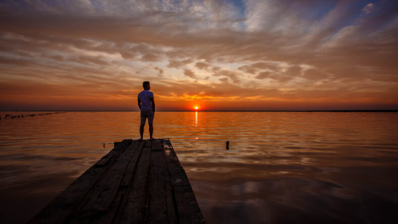 A man stands on a wooden pier and looks at the horizon of the sunset of the lake.
