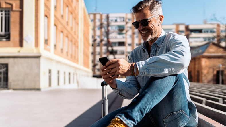 Stock photo of a business man seated in a bench in the street looking at his mobile phone and listening to music with his wireless earphones. He is holding his smartphone with his two hands.
