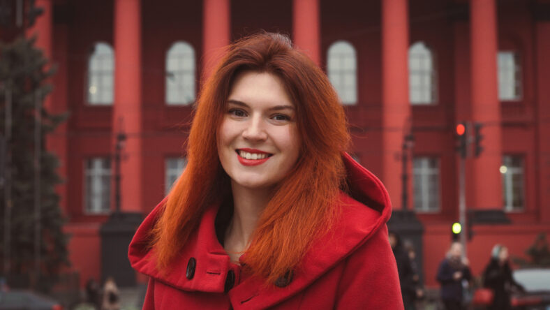 Close up young smiling woman in red coat on street portrait picture. Closeup front view photography with public building on background. High quality photo for ads, travel blog, magazine, article