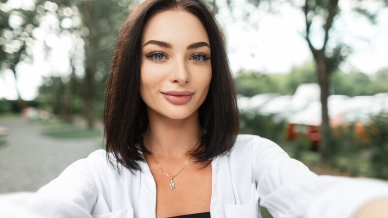 Beautiful happy woman with a smile in a fashion white shirt walking in the park and taking selfies on the phone. Female portrait of pretty girl