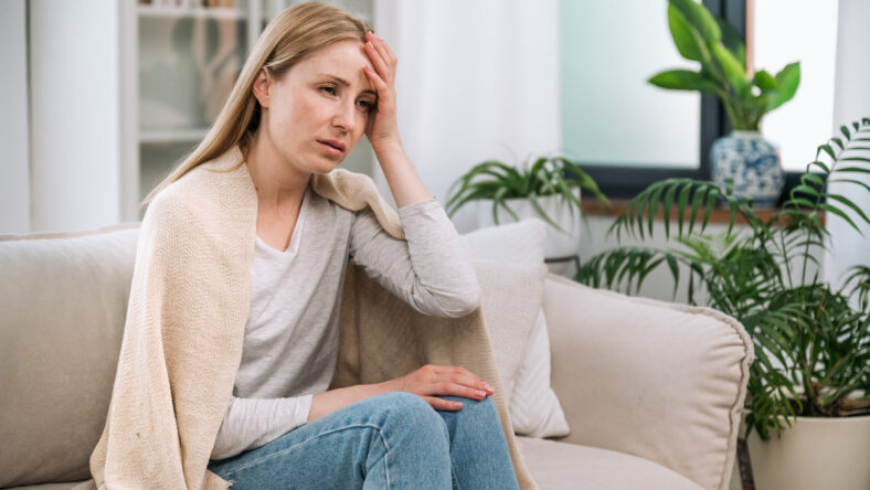 Sick woman look tired, sitting alone on couch, feeling cold and covered with warm plaid in the living room. Seasonal issues, virus diseases, having flu, headache, migraine concepts