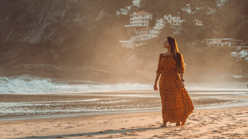 Beautiful brunette girl walking barefoot at the beach wearing a dress by sunset time