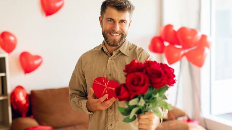 Romantic European man holding beautiful bouquet of red roses and gift box on valentines day, saying I love you, stretching flowers to camera