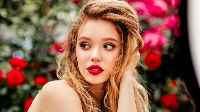 A young woman with red lips posing in a garden of roses, her expression reflecting beauty, joy, and style.