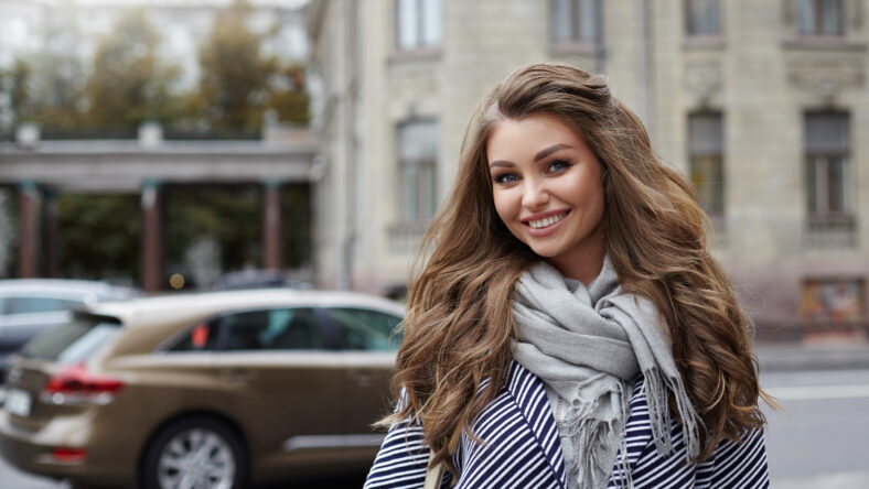 Beautiful cheerful young lady with voluminous hair wearing stylish coat and scarf posing on city street on her way to store, having joyful happy look, enjoying warm spring day. People and lifestyle