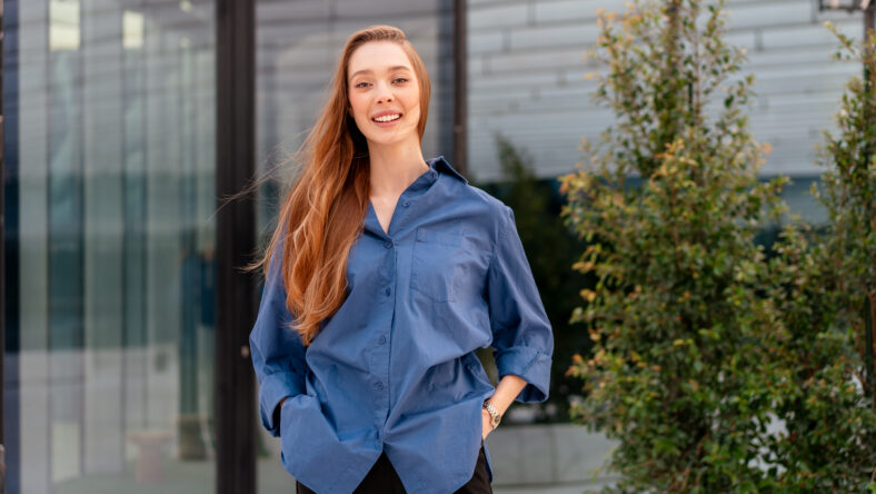 Smiling businesswoman in blue shirt standing near modern glass office building with confident and positive expression during daytime