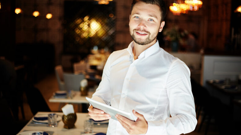 Cheerful elegant man in white shirt holding tablet and smiling at camera standing in modern restaurant