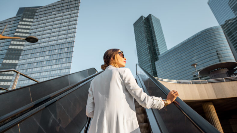 Stylish businesswoman in white suit going up on the escalator at the business centre outdoors with skyscrapers on the background in Paris