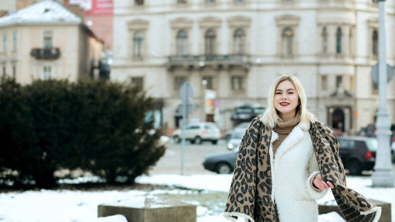 Cheerful blonde woman wears winter coat and knit scarf walking at the city. Space for text