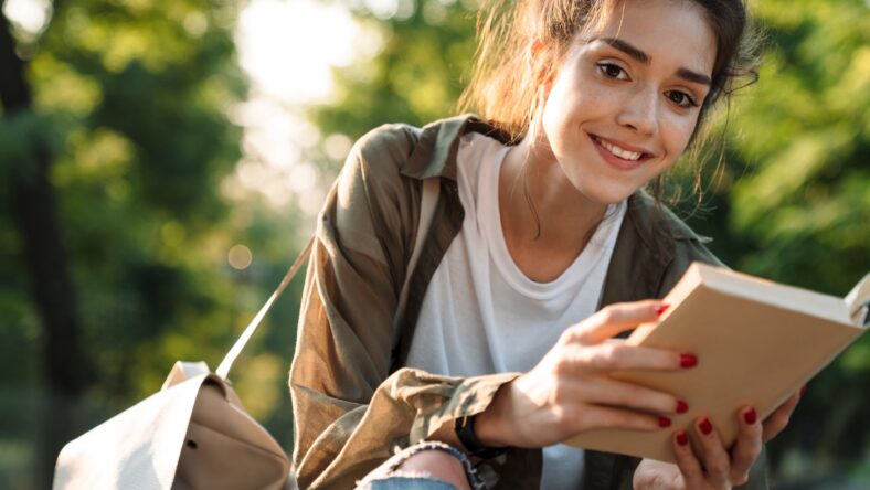 Image of caucasian woman wearing casual clothes smiling and reading book while walking in green park