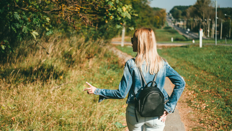 Slender blonde girl, in a blue denim jacket and light jeans and with a black leather backpack walks down the road against the backdrop of the autumn forest and highway. View from behind. Her fingers show a sign of victory.