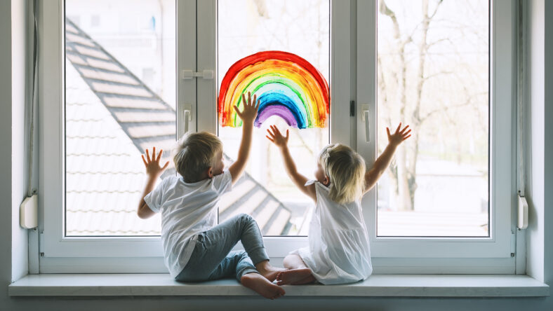 Little children on background of painting rainbow on window. Photo of kids leisure at home. Positive visual support during quarantine Pandemic Coronavirus Covid-19 at home. Family background