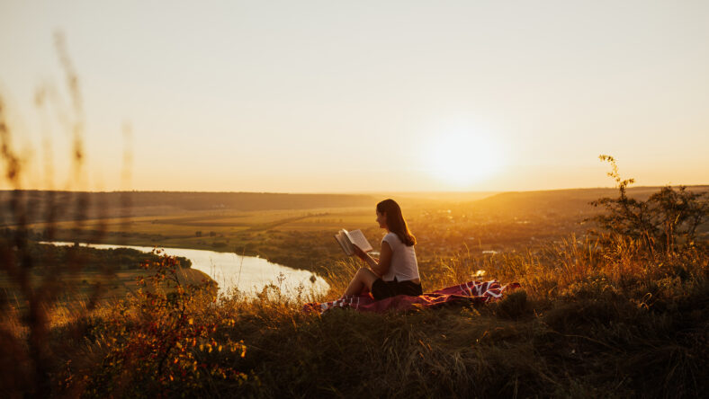 Calm girl reading a book on hill with perfect landscape, enjoying time on holiday. She read book in quiet nature.