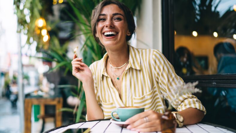 Positive young female in casual clothes smiling and drinking cup of coffee and holding teaspoon while spending time in outdoor cafeteria