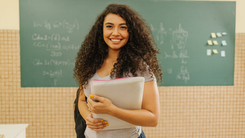 Latin Curly haired student smiling wearing backpack holding a notebook in a classroom