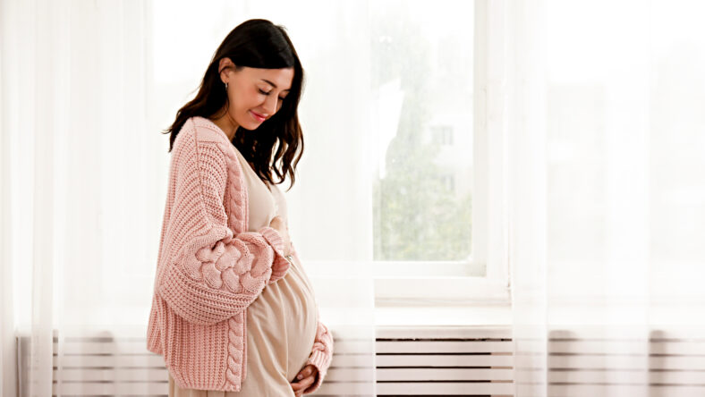 Young beautiful woman on second trimester of pregnancy. Close up of pregnant female in pink oversized knitted sweater with arms on her round belly. Expecting a child concept. Background, copy space.