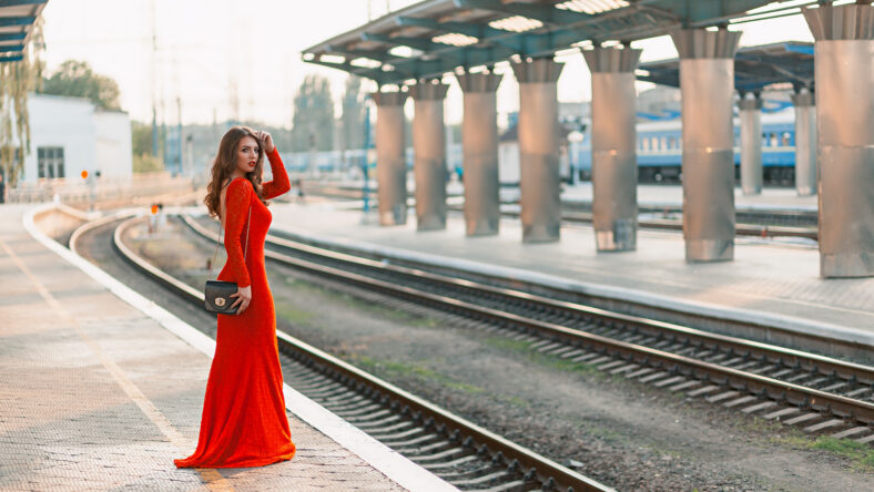 beautiful young woman in a long red dress stands on the platform of the station near the railway.