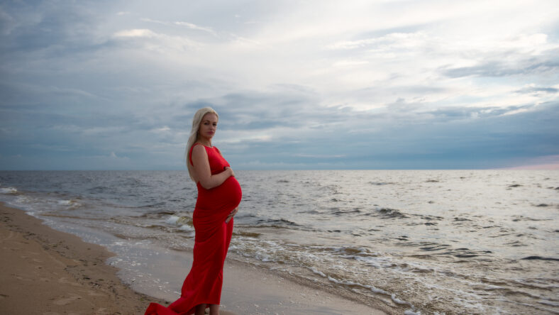 Beautiful pregnant woman in red dress standing at the beach during sunset