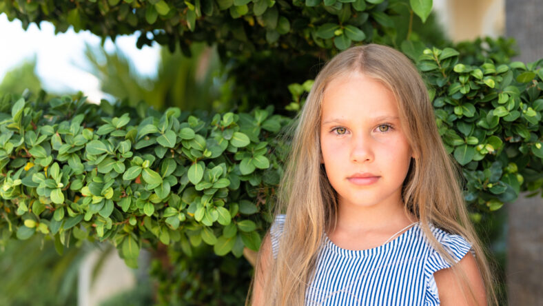 portrait face of candid happy little kid girl of eight years old with long blond hair and green eyes on background of green plants during a summer vacation travel. gen z mental health concept
