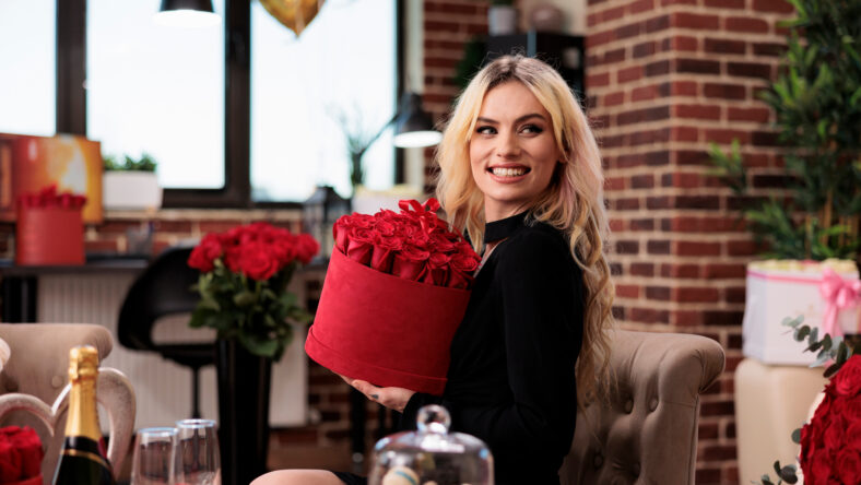 Smiling beautiful woman holding valentines day red roses box, looking at camera. Attractive blonde girlfriend sitting in living room with expensive gifts, receiving romantic luxury presents
