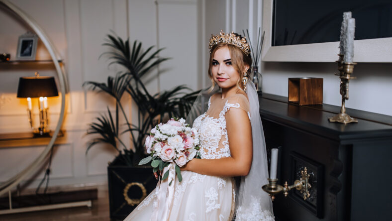 A beautiful young blonde bride with a crown, a diadem in a white lace dress sits indoors, a cafe near an old piano with a bouquet in her hands. Wedding photography, close-up portrait, vintage.