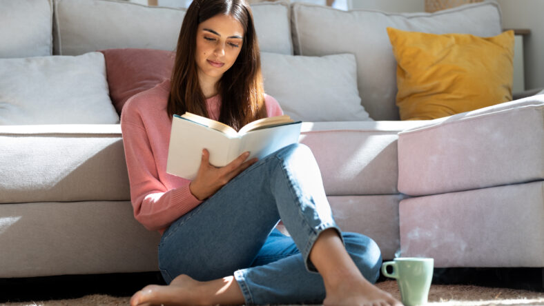 Shot of pretty young woman reading a book and drinking a cup of coffee while sitting on the floor at home.