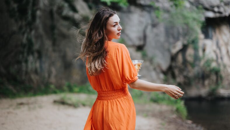Woman in an orange dress holding a glass of champagne, enjoying a picnic in a serene outdoor setting by the water.