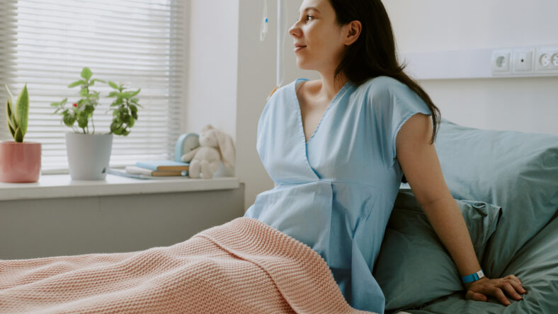 Young pregnant woman sitting on bed and looking out of window while she staying at maternity hospital