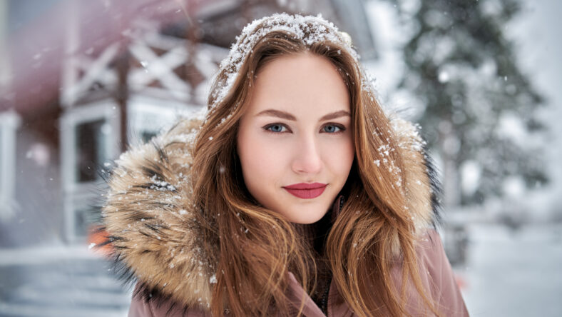 Winter portrait of a beautiful young woman in a warm jacket with fur hood against the backdrop of a snowy landscape.