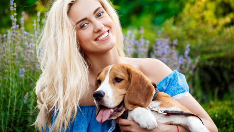 Young beautiful blonde girl in blue dress smiling, walking, playing with beagle dog in park.