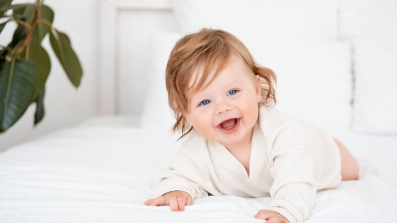 A baby with red hair and blue eyes in a white bodysuit is smiling happily on the bed at home, a healthy happy cute newborn baby boy.