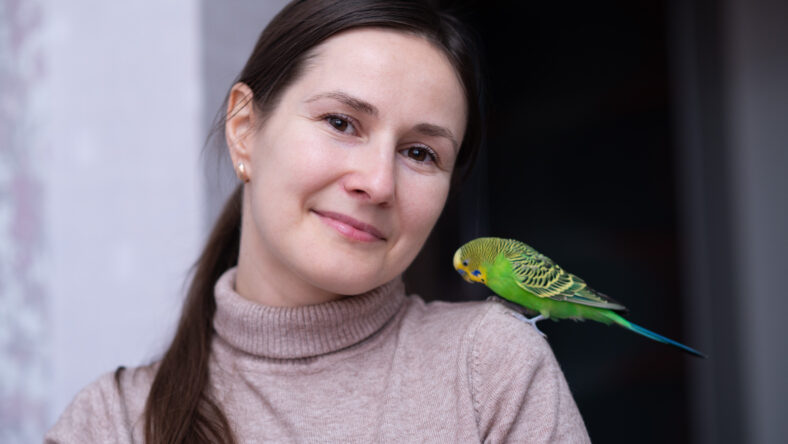 smiling brunette woman in a beige turtleneck with a bright green budgie perched on her shoulder, dark background