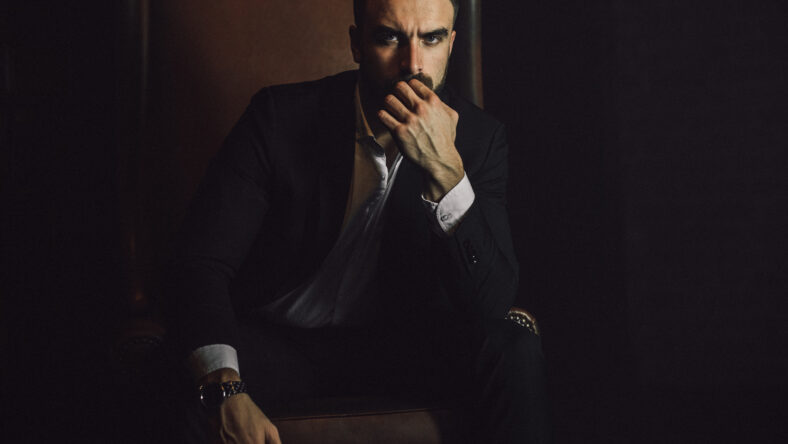 A handsome man with a beard and in a classic suit sits in a leather chair and looks at the camera.