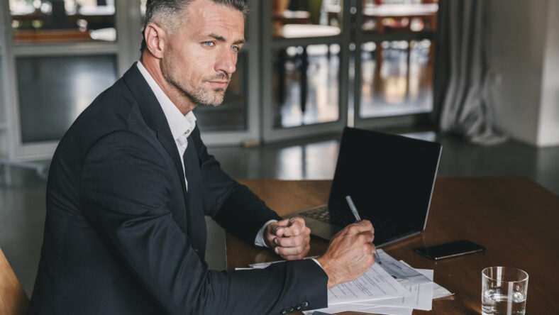 Image of concentrated confident businessman 30s wearing white shirt and black suit sitting at table in office during work with documents and laptop