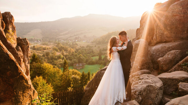 Romantic wedding couple of groom and bride hugging and kissing while standing on the top of the cliff in national park with beautiful view on sunny summer day. Sun rays