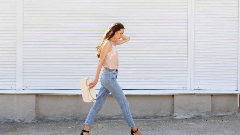 Young stylish woman wearing beige cami silk top, blue cropped denim jeans, black high heel sandals and holding clutch bag walking in the city street. Trendy casual outfit. Street fashion.