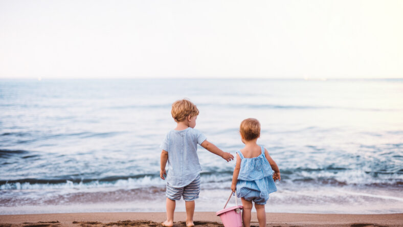 A rear view of two toddler children playing on sand beach on summer family holiday.