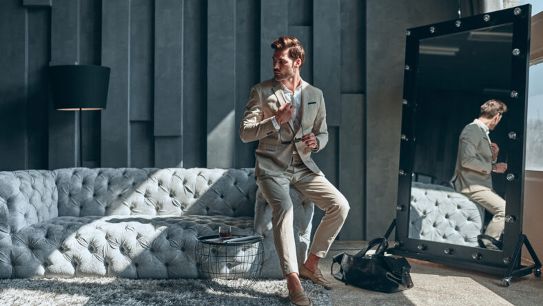 Handsome stylish man in beige suit at home sitting on sofa near mirror.