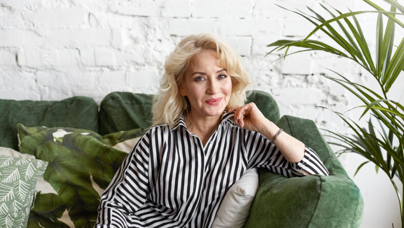 People, joy, positiveness, lifestyle and age. Horizontal shot of beautiful joyful mature female wearing elegant clothes enjoying leisure time at home, looking and smiling mysteriously at camera