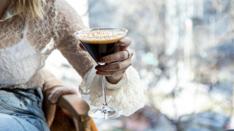 close up view of a woman's hand picking a martini glass filled with delicious chocolate coffee martini with sprinkles. blurred view of the city buildings with sunglasses and black hat on coffee table