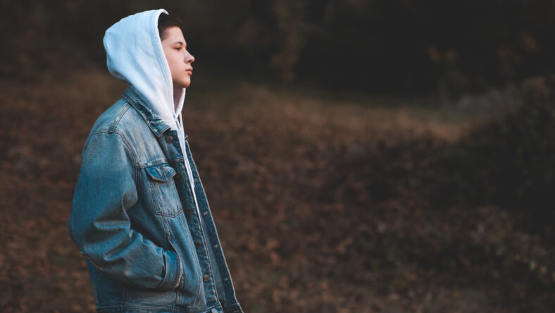Handsome teen boy 15-16 year old wearing denim jacket and white hoodie standing outdoors over nature autumn background. Teenagerhood.
