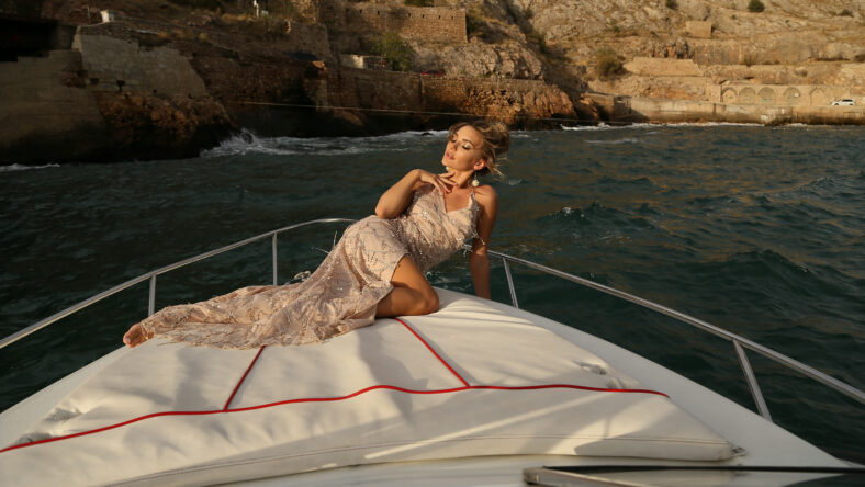 beautiful woman with blond hair in luxurious dress posing in boat with antique buildings on background