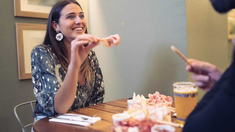Couple sitting in a restaurant table having fun talking and joking together eating aperitif with bread stick, salami and drinking beers