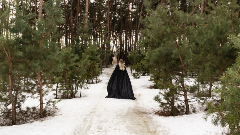 Blonde woman in a black cloak with a hood in winter in a pine forest. Female in cloak from the view from the back side