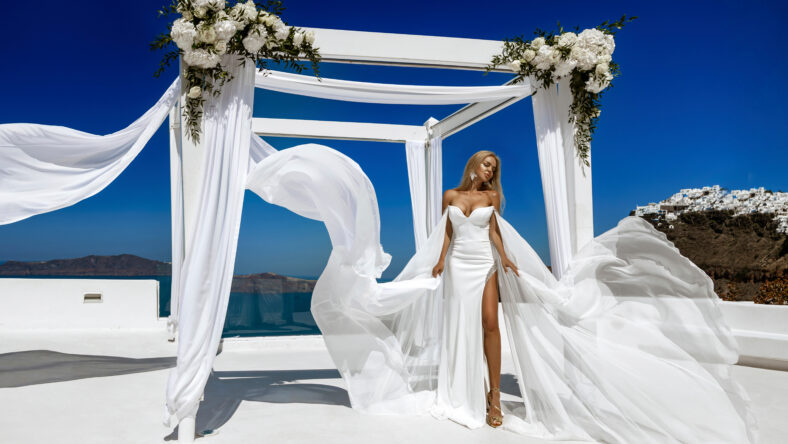 Stylish elegant bride woman in white long fluttering wedding dress is posing in wedding area in Fira in Santorini.