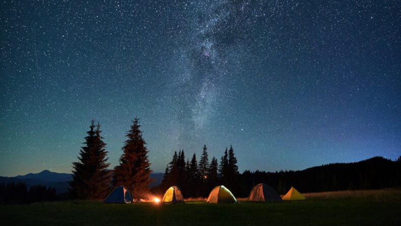 Night camping in mountains under starry sky. Tourist tents in campsite near burning campfire under beautiful sky full of stars with Milky way above forest. Concept of tourism and traveling.
