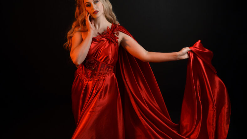 Close up portrait of beautiful blonde model wearing flowing red silk toga gown and crown, dressed as ancient mythological fantasy goddess. Graceful elegant pose isolated on dark studio background.