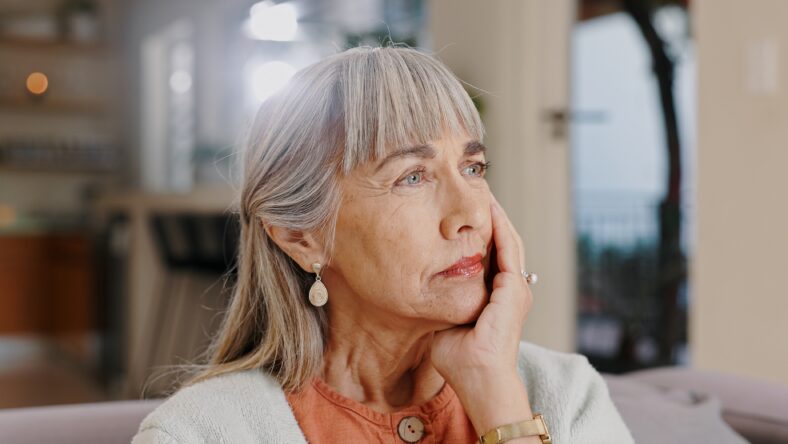 Senior woman, thinking and sad in lounge, hand and memory of past, retirement and depression in nursing home. Grandma, lonely and couch of house, remember and elderly person in living room and grief.