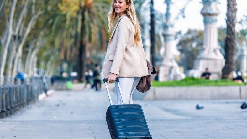 Shot of pretty young woman walking with suitcase while looking at camera through the city