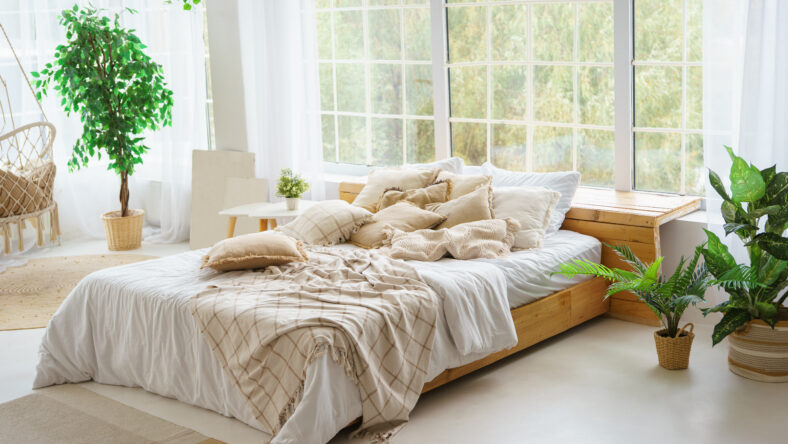 Comfortable bed with lots of soft decorative beige pillows in light sunny bedroom with green plants and large window. Interior design of living room in boho style